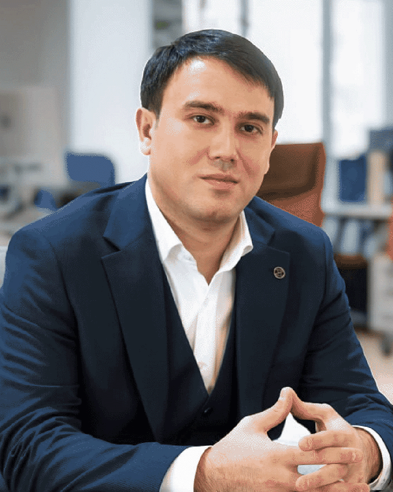 Professional portrait of Uzbek man in his 50s with graying hair wearing dark business suit in modern corporate office setting